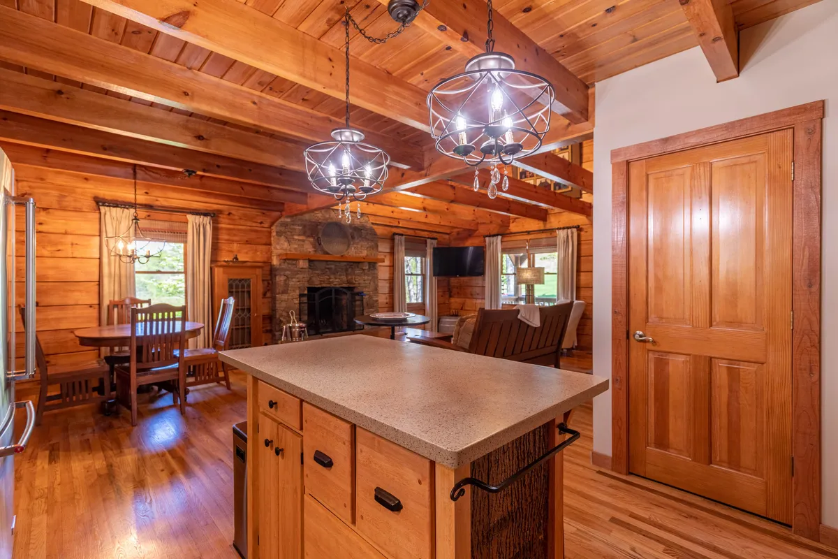 A spacious, inviting open-plan living area featuring a rustic wooden dining table set near a beautiful stone fireplace. The setting is brightened by ample natural light streaming in from large windows, while comfortable furnishings and a warm color palette create a welcoming atmosphere. The presence of the fireplace and the sturdy table evoke feelings of togetherness and warmth—ideal for family gatherings or post-adventure relaxation.