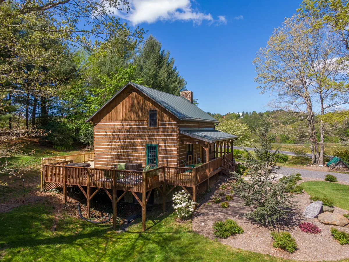 The image displays a spacious wooden deck attached to a log cabin, surrounded by lush greenery and flowering shrubs. The outdoor area is set up for dining and relaxation, offering views of nature and a tranquil atmosphere ideal for unwinding after a day of exploring downtown or the nearby Blue Ridge Parkway.