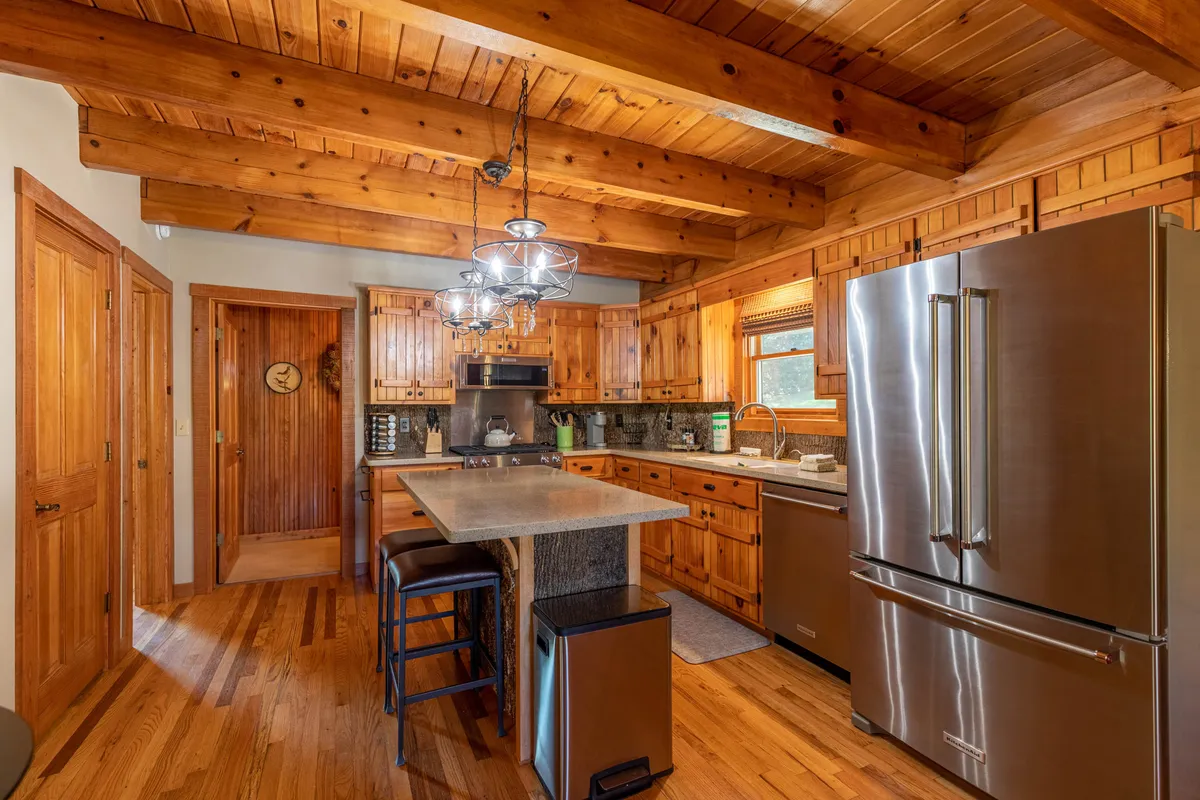 This image highlights a spacious kitchen anchored by a central island bar with seating, surrounded by rich wooden cabinetry and exposed beams. Stainless steel KitchenAid appliances, including a prominent gas range, are visible. The setting is warmly lit and welcoming, evoking a place for shared meals and celebrations.