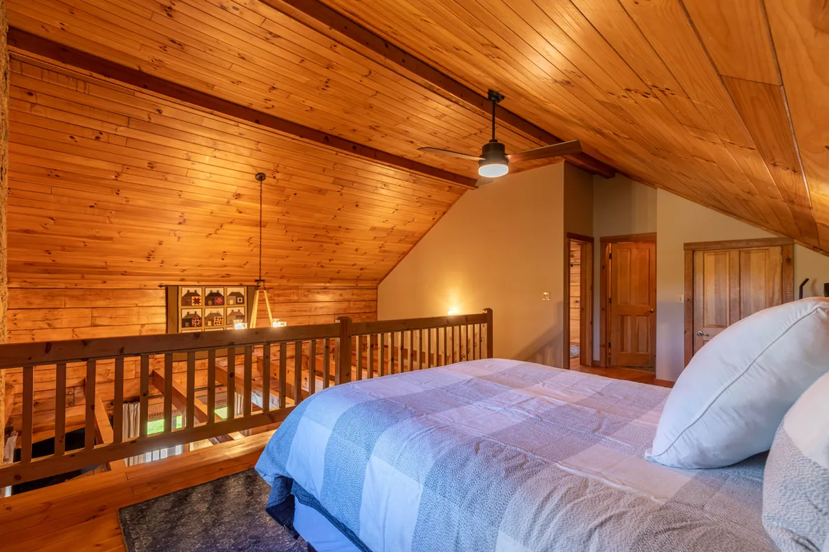 A serene loft space featuring a bed with plush decorative pillows, a wooden ceiling, and a soft lighting fixture. The loft's open layout is complemented by natural light and a wooden railing, evoking a sense of togetherness and shared retreat.