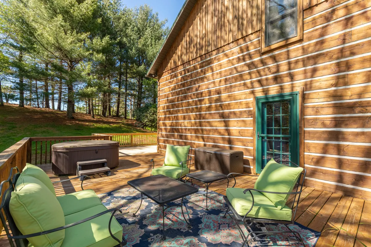 A cozy wooden deck adjacent to the house, featuring a hot tub, green seating, and a sliding door that connects indoor and outdoor living. The setting is ideal for stargazing, morning coffee, and fireside gatherings, with plenty of space for pets and family activities.