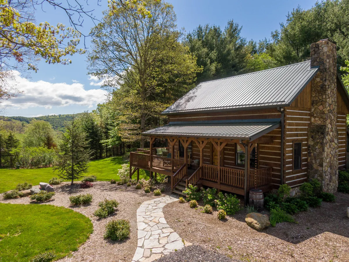 The image showcases a charming log cabin with a classic stone chimney, a welcoming front porch, and a beautifully landscaped garden set against a peaceful mountain backdrop. The porch is positioned for enjoying stunning sunsets, morning coffee in the crisp mountain air, and evenings under the stars, all surrounded by lush nature.