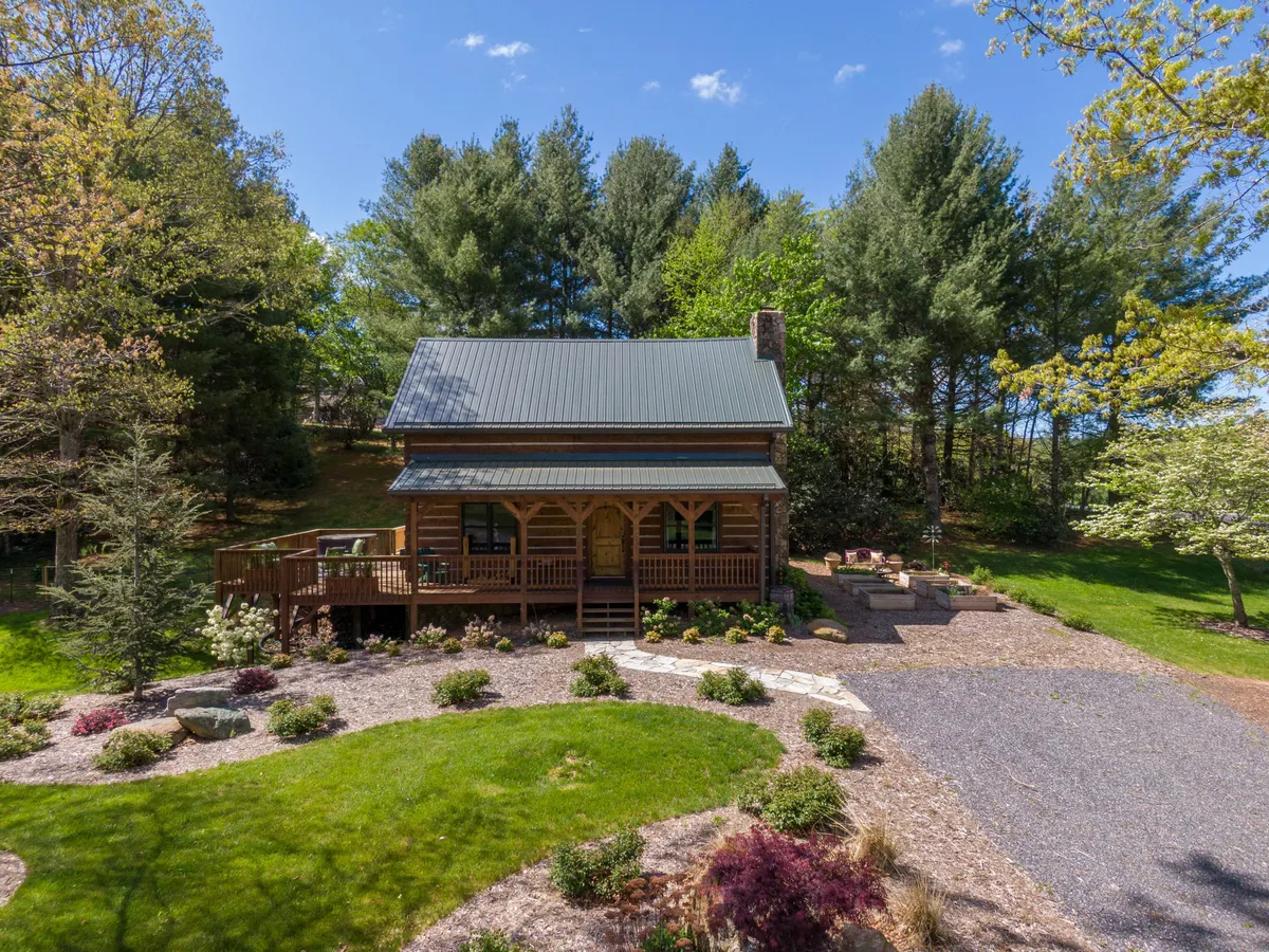 A charming log cabin with a spacious wrap-around porch, nestled amid lush greenery and a landscaped garden. The scene exudes serenity, inviting guests to relax on the porch, enjoy the peaceful natural surroundings, or gather around a fire pit in the evening. The cabin’s exterior highlights its rustic appeal and connection to the mountain environment.
