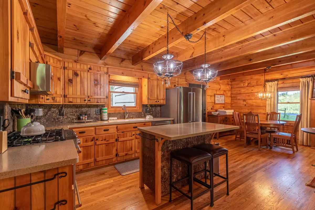 A cozy view of the kitchen and adjoining dining area, featuring wooden cabinetry, a centrally placed island with seating for casual breakfasts, and a rustic dining table under warm lighting. The inviting layout encourages family gatherings and shared culinary adventures.