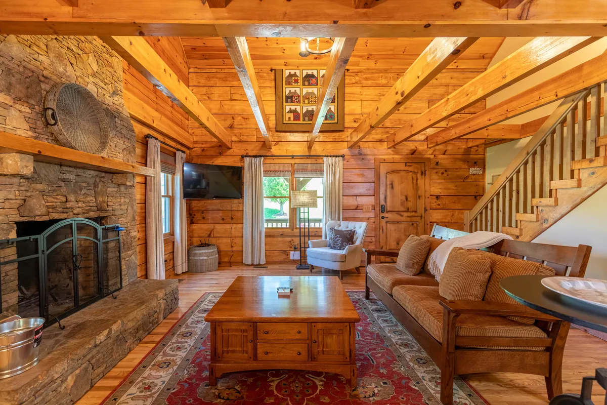 A cozy living space centered around a stone fireplace, with exposed wood beams overhead, a comfortable sofa, a coffee table, and stairs leading to another level. Natural light pours in, enhancing the rustic charm and inviting ambiance ideal for family gatherings and relaxation.