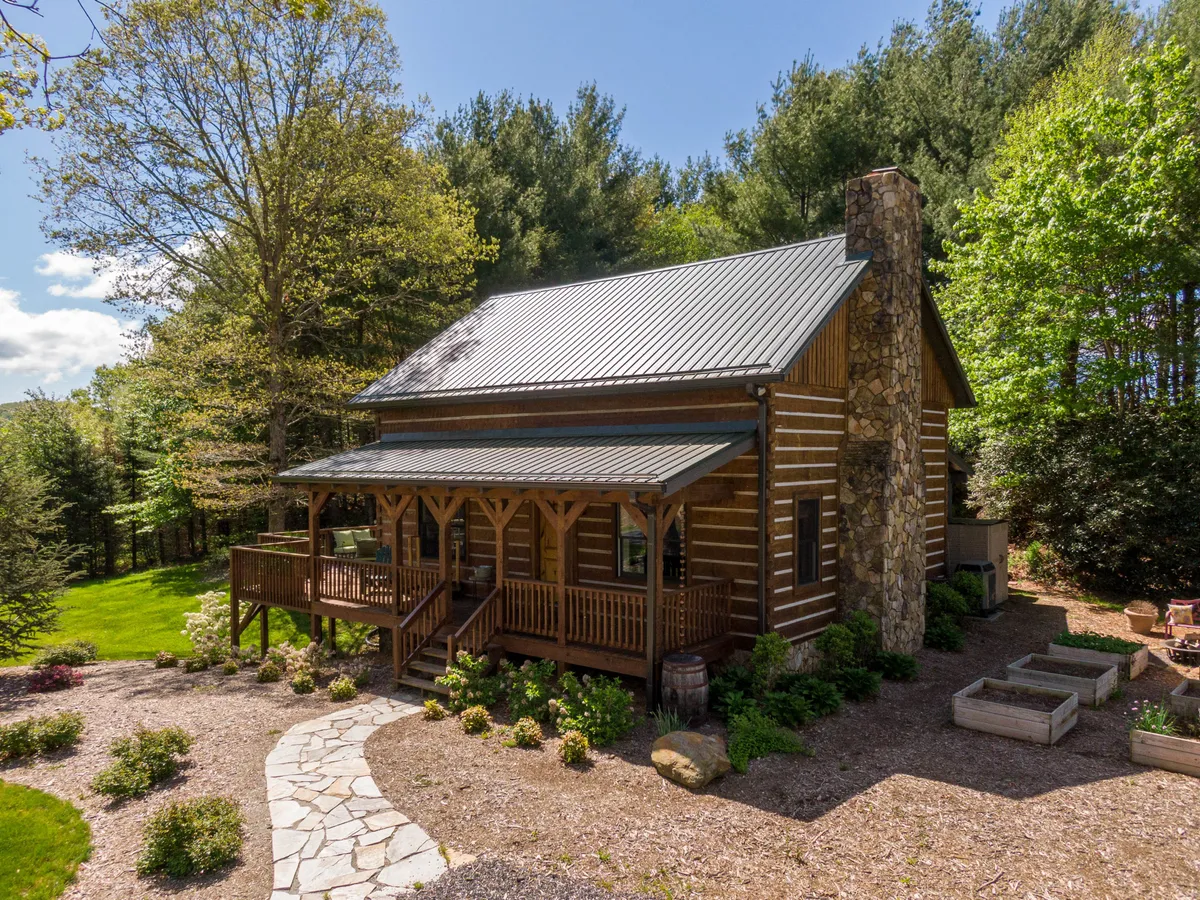 A picturesque log cabin featuring a stone chimney and front porch, surrounded by a landscaped yard and lush greenery. The inviting exterior hints at cozy moments inside and provides a welcoming sense of arrival after a day exploring the mountains.