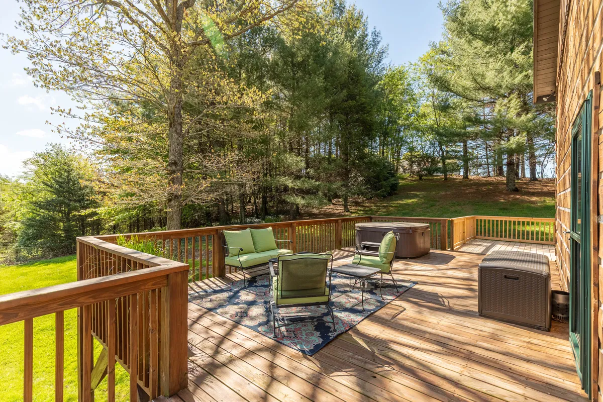 A spacious wooden deck set against a lush backdrop of greenery, featuring a comfortable seating area, a hot tub, and a grill. The inviting layout makes it perfect for relaxing, dining, or entertaining outdoors, while the surrounding nature provides privacy and tranquility.