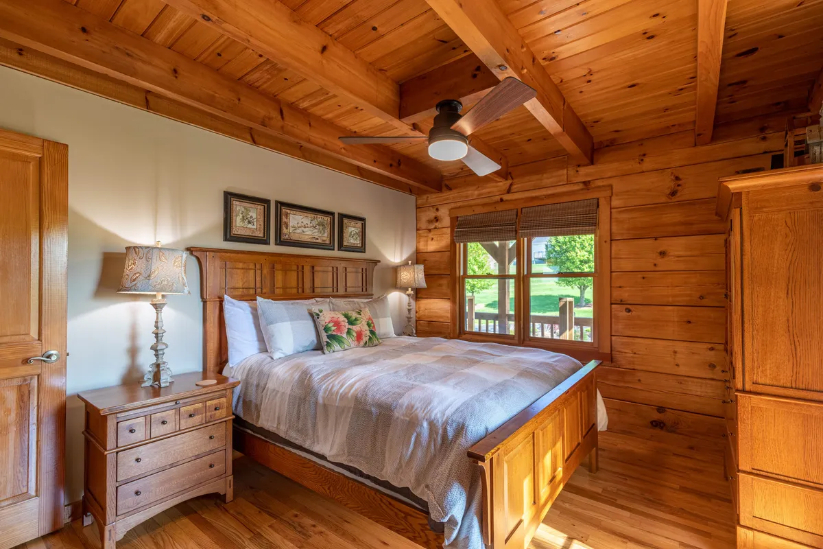 A warmly lit bedroom with a wooden bed frame adorned with decorative pillows, flanked by lamps and framed artwork. Natural light streams in through the window, creating a peaceful and inviting atmosphere ideal for restful nights.